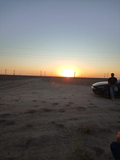 In Haratan, Uzbekistan, at sunset, a man in a black t-shirt, blue jeans, and sunglasses leans against a dark-colored car parked on a dirt road. The sun is setting on the horizon, casting a warm glow across the sky, which transitions from a pale blue at the top to a vibrant orange and yellow near the horizon. Power lines stretch across the background, supported by poles. The terrain is dry and dusty, with sparse vegetation.