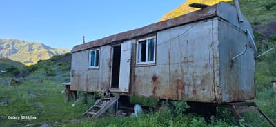 This media file depicts a rustic, weathered mobile home or trailer situated in a natural, mountainous landscape. The structure is metallic, showing significant signs of rust and age, with a curved roofline. It has two windows with white frames and a single doorway that is open, revealing a dark interior. A makeshift wooden staircase leads up to the entrance.

The trailer is positioned on uneven ground, surrounded by tall green grass and scattered blue wildflowers, suggesting a rural or remote setting. In the background, rolling hills and mountains are visible, bathed in the warm light of what appears to be late afternoon or early evening. The sky is clear and blue.

Notable details include a satellite dish mounted on the side of the trailer, a chimney pipe protruding from the roof, and some electrical wires running along the exterior. Some discarded items, including a white plastic container and possibly barrels, are visible near the base of the trailer. The overall impression is one of isolation and a somewhat neglected but functional dwelling.

The visible text at the bottom left of the image reads "Galaxy S24 Ultra," indicating the device used to capture the photograph. There are no people visible in the scene. The location context provided is Almaluu, Kyrgyzstan, which aligns with the mountainous terrain and the rural, solitary feel of the image. The time of day is likely late afternoon due to the golden sunlight illuminating the hills. The weather appears to be clear and pleasant.