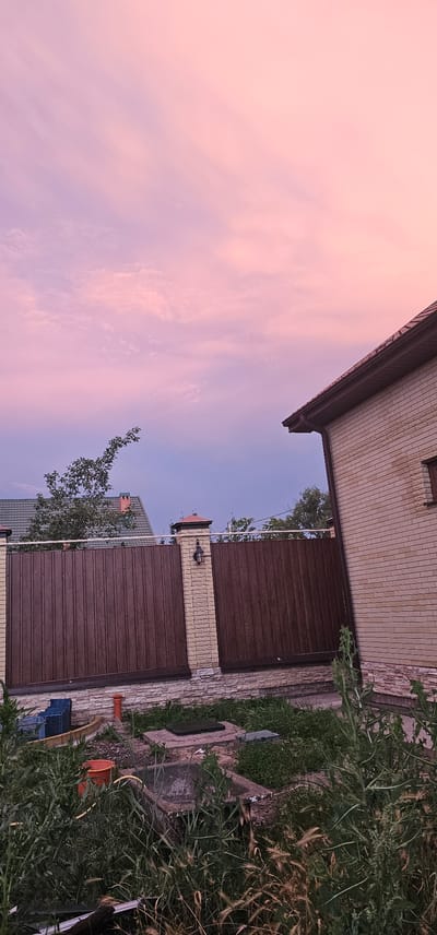 The image depicts a suburban outdoor scene, likely at dusk, with a focus on a fence, a house, and the sky. The sky is filled with soft, pastel-colored clouds, predominantly in shades of pink and purple, indicating either sunrise or sunset.
In the foreground, a wooden fence with vertical planks is visible, supported by brick pillars. A decorative lantern is mounted on one of the pillars. To the left of the fence, a green-tiled roof of another building is partially seen, along with the branches of a leafy tree. A portion of a light-colored brick house with a brown-tiled roof is to the right, extending towards the edge of the frame.
The ground in front of the fence is unkempt, with overgrown grass and several concrete structures, possibly manholes or water features. Scattered around are some plastic crates, an orange bucket, and a yellow hose.
The overall impression is a quiet, residential setting under a visually striking sky. There are no people present in the image. The location is identified as within the city of Овощной, Russia.