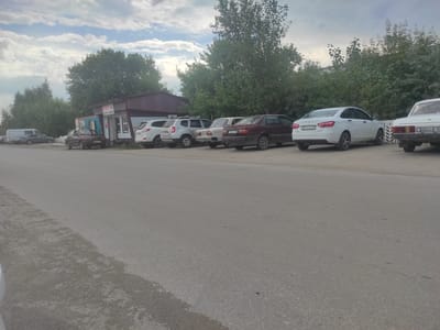 This image shows a row of parked cars on the side of a paved road in front of a small building with a maroon roof. The building appears to be a small shop or kiosk with signs in Cyrillic script. The cars are various models and colors, including a white sedan with a license plate that reads "E554XH 10". Trees and foliage line the background, and the sky is cloudy. The scene is likely located in Denisovka, Kazakhstan, given the Cyrillic script and the general appearance of the environment. There are no people visible in the image.