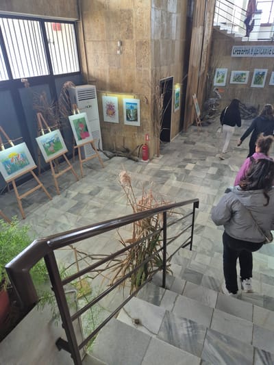 The image shows an indoor exhibition space in Srebarna, Bulgaria. Several paintings are displayed on easels and mounted on the wall, featuring themes such as landscapes and animals. There are visitors viewing the exhibition. The space has a tiled floor, and a staircase with a metal railing is present. A fire extinguisher is mounted on the wall. There are dried floral arrangements and decorative elements. The architecture appears to be a combination of exposed stone or concrete walls and large windows with blinds.
