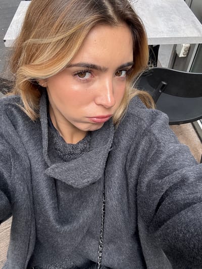 The image depicts a close-up, chest-up selfie of a single female subject, approximately 20-35 years old, in the city of Vaulx-en-Velin, France. She has light skin, long brown hair with prominent blonde highlights, parted centrally, and brown eyes looking directly into the camera. Her lips are glossy and positioned in a deliberate pout. A small, light-colored stud earring is visible in her right earlobe. She is wearing a dark grey, textured overcoat or jacket with an unstructured, possibly cowl-neck collar, layered over a darker grey, possibly knitted, garment. A thin, dark-colored chain, likely from a bag, is visible crossing her chest.
The background, partially visible on the right, shows a section of a light grey, rectangular table surface. Below the table, a dark-colored chair with a rectangular cutout in its backrest is present. An off-white electrical outlet or fixture is affixed to a vertical surface adjacent to the table. A light brown, textured floor or ground surface is visible at the bottom right. A lighter, possibly white, vertical surface is faintly discernible in the upper left background. The overall scene suggests an indoor or sheltered outdoor setting.