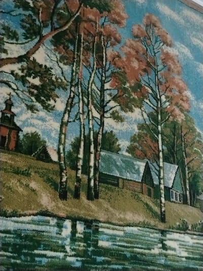 The image depicts a pastoral scene with a body of water in the foreground, reflecting the sky and trees. On the embankment beyond the water, there are several buildings with steep, dark roofs, suggesting a rural settlement. A prominent church with a dark dome and a cross is visible on the left side of the embankment, situated amidst trees. Various trees, including some with autumnal foliage, are scattered throughout the scene, with birch trees being particularly noticeable due to their distinctive striped bark. The sky is blue with white clouds. The overall impression is one of a tranquil village or countryside setting. The presence of the church and the architecture suggests a location within Russia, specifically referencing a village named Лычково.