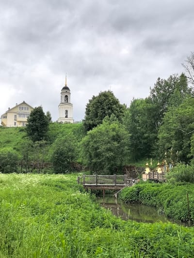 A wide shot captures a lush, green landscape with a cloudy sky overhead. In the foreground, a dense field of wild flowers and tall grasses borders a gently flowing stream. A wooden footbridge, with a railing, crosses the stream, leading to a path on the opposite bank.
On the rising hill in the background, a pale yellow building with multiple windows is situated to the left. Next to it stands a tall, white bell tower topped with a golden spire, a bell visible within its open structure. Further to the right on the hillside, a cluster of golden-domed structures, possibly chapels or smaller churches, are visible amidst the trees. The scene is dominated by abundant green foliage, with trees of various sizes forming a thick backdrop. The overall impression is one of a peaceful, rural setting in Russia. The lighting suggests it is daytime, possibly overcast. No text is visible.