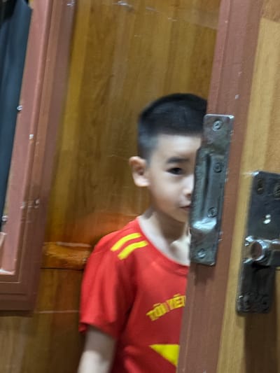 A young boy wearing a red shirt with yellow stripes on the sleeves and a yellow emblem is partially visible behind a wooden doorframe. The boy's face is turned to his left, with his right eye and part of his cheek visible. The background consists of wooden walls and a brown door. The scene is likely indoors in a residential setting within Hải Vân Ward, Vietnam.