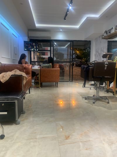 The image displays the interior of a commercial establishment, specifically a salon or beauty spa, situated in Lauro de Freitas, Brazil, observed during nighttime conditions. The floor is composed of large, polished, light-colored tiles with prominent brown and orange veining, reflecting multiple points of orange light, likely from exterior sources.
On the left side of the frame, a dark-colored mobile pedicure station with a black drawer and wheels is present, topped by a brown surface partially covered by a light-colored towel. Adjacent to this, two individuals are seated in brown, tufted armchairs. The person on the far left, a female with dark hair, is wearing a dark top and appears to have a mask on, facing right. The second individual, also a female with dark hair, wears a patterned top (possibly green/beige) and is seated in an identical armchair, positioned opposite a small, dark-colored table between the two chairs, indicating an ongoing beauty service. Above these individuals, a flat-screen television is wall-mounted, displaying an indiscernible image of what appears to be a crowd or event. An air conditioning unit is mounted directly above the television. The wall supporting the television features white paneling with a vertical strip of blue LED lighting on the left.
The central and right portions of the image reveal a large glass storefront or wall, offering a view of the exterior night scene. Outside, a dark sky is visible above a paved street. Partial views of a dark-colored vehicle and a multi-story building with red and white sections are present across the street. Streetlights contribute to the ambient illumination outside, with their light sources reflecting on the interior floor. To the right, a dark brown salon chair with a chrome base and a black footrest is positioned. Further to the right, a light-colored wooden shelving unit displaying bottles and products is situated against a white wall adorned with a grey marble pattern. The ceiling throughout the interior is white, featuring both recessed lighting and a black track light fixture equipped with multiple spotlights.