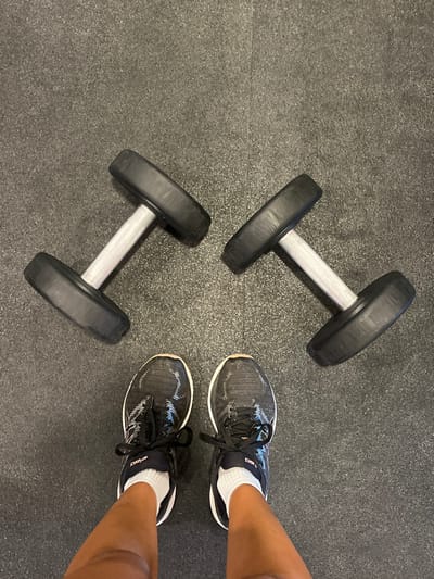 A person is standing on a black rubber mat in a gym located at 25.657, -100.449, Ciudad Santa Catarina, Mexico. Two dumbbells are lying on the ground in front of the person. The person is wearing black running shoes with white soles and laces. The person's lower legs and feet are visible in the image. The person is looking down at the dumbbells. The image is taken from a first-person perspective. The rubber mat is textured and has a slightly faded appearance. The lighting in the room is dim.