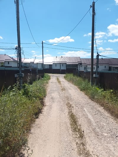 The image depicts a rural dirt road leading through a residential area. Two prominent wooden utility poles, laden with electrical wires, stand on either side of the road, framing the view towards a row of houses. The houses, appearing to be single-story with white walls and dark tiled roofs, are bordered by a dark brown, ribbed metal fence. Lush green grass and weeds grow abundantly along the sides of the road and fence. The sky is a bright blue with scattered white clouds, indicating fair weather. The scene suggests a quiet, sun-drenched day in a suburban or village setting. The location is identified as Krymsk, Russia.