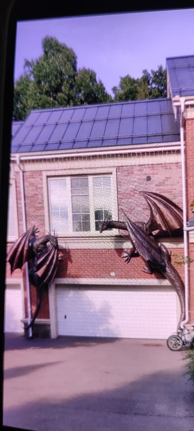 This media file depicts a house with two dragon sculptures mounted on its brick exterior wall. The house has a dark, multi-pitched roof and a large window with multiple panes. A white garage door is visible below the window. One dragon sculpture is positioned to the left, appearing to cling to the wall with its wings spread wide. The second dragon sculpture is to the right, with its body extending horizontally across the wall, and its head turned slightly towards the left. The sculptures are dark in color, with visible detailing on their wings and scales.
The scene appears to be outdoors, likely in the daytime, given the natural light illuminating the house and its surroundings. The sky is a hazy, light purple, suggesting either early morning, late evening, or perhaps a cloudy or polluted atmosphere. Trees with green foliage are visible in the background, above the roofline. A small section of a white wheeled object, possibly a bicycle or scooter, is visible in the lower right corner of the image. The ground in front of the house is paved with asphalt, and shadows are cast across it, indicating the sun's position.
The location context provided is "Московский Ветеран Снт, Russia," which translates to "Moscow Veteran Garden Community, Russia." This suggests a suburban or semi-rural setting within the Moscow region. There is no discernible text visible in the image. The overall mood is somewhat unusual due to the prominent placement of the fantastical dragon sculptures on a residential building.