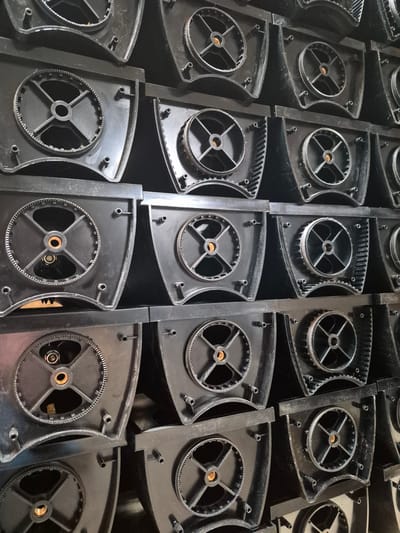 The image shows a stack of black plastic components, likely parts of an industrial machine or appliance. Each component features a circular gear mechanism with spokes radiating from a central hub. These components are arranged in a grid-like fashion, with several layers visible. The lighting is somewhat dim, and the overall impression is one of industrial storage or manufacturing. There are no people or animals in the image. The location within the city of Masów, Poland, is not discernible from the visual information provided in the image.