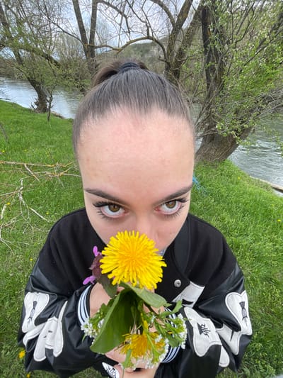 A young person with brown eyes and dark eyelashes is holding a small bouquet of dandelions and white wildflowers in front of their face. They are wearing a black bomber jacket with white designs on the sleeves. The background is a grassy area with trees and a body of water. The photograph appears to have been taken at a low angle, tilted slightly. The location is not discernible as Barkach, Bulgaria from the details in the image.