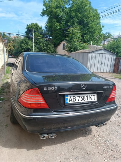 The image shows the rear passenger side of a black Mercedes-Benz S 500 sedan parked on a gravel road. The car has a license plate reading "AB 7381 KT" and the "AMG" emblem. In the background, there is a gray metal garage with the number "556" visible on it, surrounded by trees and a partially visible building. The sky is blue with some clouds. The location is identified as Mohyliv-Podilskyi, Ukraine.