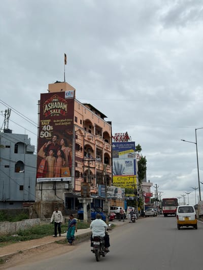 The image depicts a street scene in Shamshabad, India, under a cloudy sky. On the left side of the street, a large billboard advertises a "PRE ASHADAM SALE" with discounts of 40% and 50%, featuring an image of a family. Next to it, a multi-story building has several other advertisements and signage, including "HOTEL NEELAM," "STEEL AUTHORITY OF INDIA LIMITED," and "NEX." Further down the street, several vehicles are visible, including cars, a motorcycle with a rider, a white van, and a red bus. On the right side of the road, a street lamp stands tall. In the foreground, a dirt path runs alongside the road, with a low wall and overgrown vegetation. Two individuals are walking on the path, a man in a white shirt and dark pants, and a woman in a blue and green sari. Another person is riding a motorcycle in the foreground. The overall scene suggests a busy commercial area with a mix of buildings, businesses, and ongoing traffic.