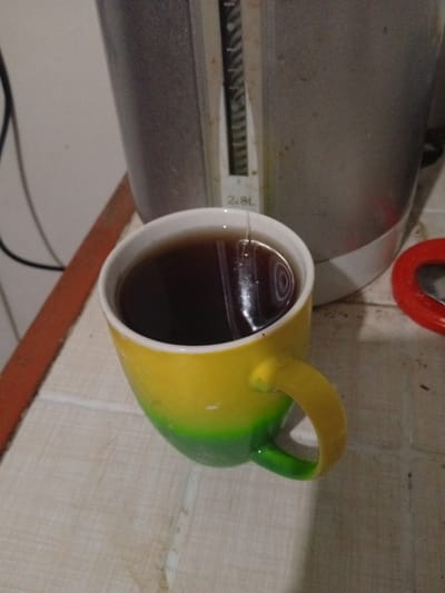 A close-up, eye-level shot shows a gradient yellow and green mug filled with dark liquid, likely tea or coffee. A tea bag string hangs into the liquid. The mug is placed on a light-colored, tiled surface. In the background, a portion of a silver and white electric kettle is visible, with a water level indicator showing "2.8L". A red object, possibly a lid, is partially seen to the right of the kettle. The environment appears to be indoors, with a wall and part of an orange-colored object visible on the left. The lighting is dim, casting shadows.