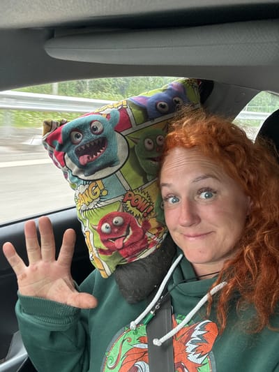 A woman with bright red, curly hair is sitting in the passenger seat of a car, taking a selfie. She is wearing a dark green hooded sweatshirt with a graphic design. To her left, propped up against the window and possibly her head, is a pillow with a cartoon monster print. The pillow features multiple colorful, cartoonish monsters with big eyes and mouths, some with comic book-style sound effects like "BANG!" and "POW!". The woman is smiling and holding her left hand up, palm facing the camera, with her fingers spread. The background visible through the car window shows greenery and a light-colored railing, suggesting an outdoor setting, possibly a road. The interior of the car, including the ceiling and a portion of the dashboard, is also visible. The lighting suggests it is daytime. The image does not provide any information about the city of Можга, Russia.