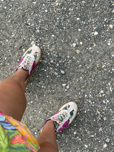 A first-person, overhead view shows a person's legs and feet standing on a gravel surface. The person is wearing tan sneakers with a tropical leaf pattern and pink laces. The person is also wearing a colorful, floral-patterned skirt. The gravel surface is a mixture of small, grey and white stones. The scene is outdoors, with no buildings or landmarks visible. The location is identified as Novalaise, France.