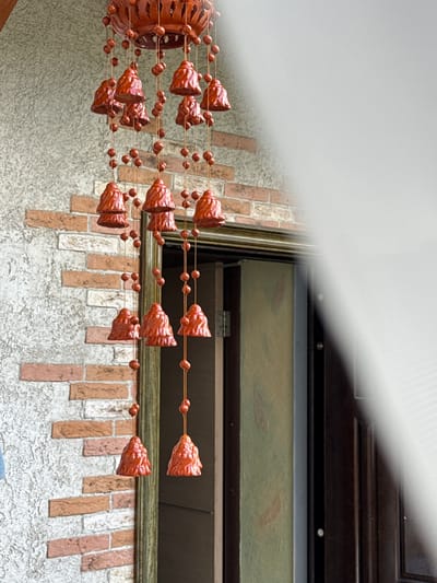 An outdoor shot focuses on a wind chime with multiple tiers of red, bell-shaped ornaments hanging from strings adorned with small, round beads. The wind chime is positioned in front of a wall constructed with textured, light-colored stucco and a decorative pattern of reddish-brown bricks. To the right of the wind chime, a portion of a doorway is visible, featuring a dark wooden frame and what appears to be a glass pane with a faint, abstract design. The overall scene suggests a residential exterior in Bersenevka, Russia, likely on a building facade.