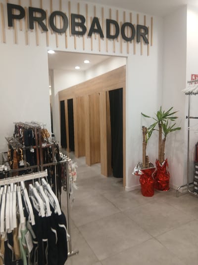 The image shows the interior of a clothing store in Ávila, Spain. The scene is brightly lit, suggesting it is daytime. Dominating the left side of the image is large, dark lettering spelling "PROBADOR" which translates to "fitting room" in Spanish. These letters are mounted on a white wall with horizontal wooden slats. To the right of the "PROBADOR" sign, there is a wooden structure that appears to be a fitting room or changing area. In front of this structure, and slightly to the right, are two potted plants with green leaves, wrapped in bright red and silver foil.
Further to the right, a metal rack is filled with neatly folded clothes, mostly dark-colored tops and sweaters, displayed on white hangers. The clothing is arranged in multiple rows, suggesting a retail environment stocked with merchandise. In the background, a tiled floor is visible. At the top of the image, a red fire extinguisher sign is mounted on the white wall. The overall impression is of a clean, modern retail space. There are no people visible in the image.