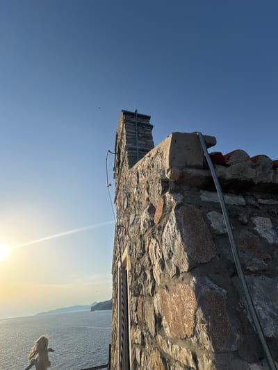 In Brežine, Montenegro, a stone building with a chimney stands against a clear blue sky. The sun shines brightly, casting a lens flare across the scene and illuminating the ocean in the distance. A young girl with blonde hair is visible on a balcony, leaning over a railing. The building's facade is constructed from rough-hewn stones of varying sizes and colors, with some reddish-brown stones interspersed among gray and white ones. A metal pipe runs vertically along the side of the building. The ocean stretches out towards a hazy coastline with several islands or peninsulas. The overall impression is one of a warm, sunny day by the sea.