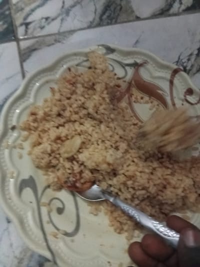 The image shows a close-up view of a plate of food, likely jollof rice, in Pantinapu, Nigeria. A hand is visible on the right side of the frame, holding a silver spoon which is partially submerged in the rice. The plate itself is decorative, with a patterned rim featuring swirls and brown accents. The rice appears to be cooked with tomato paste, giving it a reddish-brown hue. Some small pieces of what might be meat or vegetables are interspersed within the rice. The background is blurred, but suggests a tiled surface. The lighting appears to be natural, possibly from daytime, but the overall image quality is low, making it difficult to ascertain finer details. The focus is entirely on the food and the action of eating.