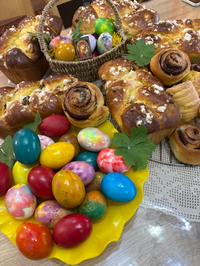 The image shows a display of Easter foods. A yellow plate is filled with brightly colored Easter eggs in shades of blue, red, yellow, green, and marbled patterns. Adjacent to the plate is a basket containing additional decorated eggs, along with an icon depicting Mary and Jesus. Surrounding the eggs and basket are various sweet breads, including braided loaves and cinnamon rolls, some adorned with crystalized sugar. Green leaves are interspersed among the food items. The scene is set indoors, likely in a home, and the location can be inferred from the context provided to be Makariopolsko, Bulgaria, given the implied connection between the image and place.