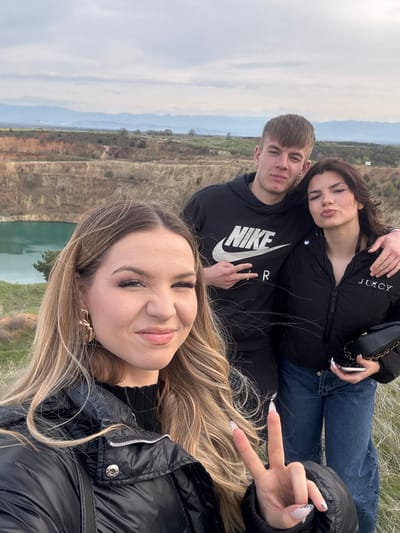 A selfie of three young people, taken outdoors in Tsar Asen, Bulgaria. The foreground shows a young woman with blonde hair wearing a black puffer jacket, holding up a hand with two fingers in a "peace" sign. Behind her and slightly to the left is a young man in a black Nike hoodie giving a peace sign as well, standing next to a young woman in a black Juicy puffer jacket with her hand on the young man's shoulder. In the background is a lake bordered by trees, below a cliff face and hills. The sky is overcast.