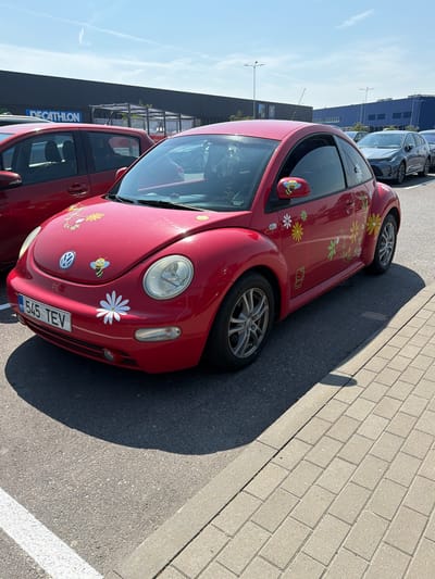 In Kurna, Estonia, on a sunny day, a red Volkswagen Beetle is parked in an outdoor parking lot. The car is decorated with flower decals and a bee sticker on the driver's side mirror. The license plate reads "545 TEV". In the background, a large building with "DECATHLON" signage is visible. Other cars are parked nearby, and the ground is paved with asphalt and interlocking bricks.