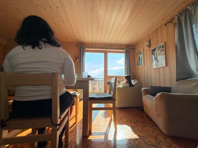 This interior scene depicts a room in Bormio, Italy, characterized by light-colored wooden paneling on the walls and ceiling, and a patterned wooden parquet floor. Bright sunlight streams through a large window and an adjacent glass door unit at the rear of the room, casting distinct rectangular shadows onto the floor.
Two individuals are present. In the foreground-left, a person with dark, wavy hair, wearing a white long-sleeved top and dark pants, is seated with their back to the camera on a light-colored wooden chair at a matching wooden table. They are oriented towards the large window, which showcases a clear view of distant mountains under a blue sky with scattered white clouds.
To the right, a second person, with dark hair tied back, is seated in a light-colored, soft armchair or beanbag chair. This individual is angled towards the window and is actively engaged with a smartphone held in both hands.
Additional furniture includes a second wooden chair with a dark cushion partially visible at the table. To the right, a larger, ribbed, beige corduroy-style sofa or armchair is situated, with a dark laptop placed on its seat or armrest. The right wall is decorated with two framed art prints or posters and features a wall-mounted light fixture. Grey fabric curtains are drawn to the sides of the window and door unit.