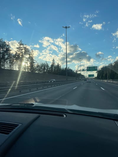 The image captures a perspective from inside a vehicle traveling on a multi-lane highway. The highway stretches into the distance, bordered by a concrete barrier on the left and a tree line on the right. Tall light poles are visible along the roadside. Several cars are driving on the highway in the same direction as the viewer. Overhead signs are present, one of which indicates "ПУНКТ ОПЛАТЫ ПРОЕЗДА" which translates to "Toll Payment Point" and a distance of "900 M". The sky is bright blue with scattered white clouds, and the sun is shining, casting rays through the trees and creating lens flare on the windshield. The foreground shows the dashboard and windshield of the vehicle. The scene appears to be set in Pesochny, Russia, given the Cyrillic text on the signage.