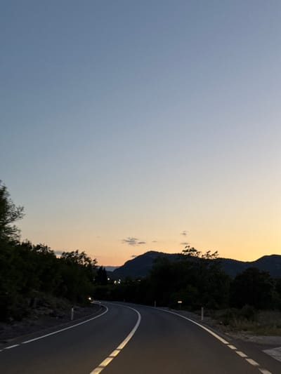 A dusky view captured from a curving road in Liješnje, Montenegro. The majority of the left side of the frame is dominated by a soft, gradient sky, transitioning from a deep blue at the bottom to a warm, sunset hue above the distant mountains. Along the right side of the image, the asphalt road curves sharply. A white line designates the road's edge, emphasizing its curve and indicating a change in direction. Adjacent to the road, a dense line of dark trees and vegetation forms a natural border, obscuring the view beyond. In the distance, the outline of mountains are visible against the bright sky, contributing to the depth and perspective of the shot. The scene is devoid of people, focusing on the tranquil beauty of the natural landscape and the engineered path through it. Illumination suggests the presence of lights along the road.