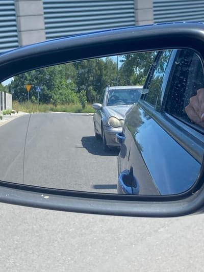 The media file shows a reflection in a car's side mirror. The primary subject is a silver car, viewed from the side and slightly from the rear, as seen in the mirror's reflection. The car appears to be parked or moving slowly on a road.

In the reflection, we can see the road surface, which is paved and appears to be asphalt. To the left of the road, there is a line of lush green trees and foliage, suggesting an outdoor, possibly rural or suburban setting. In the distance, there are two road signs visible: a yellow triangular sign and a blue square sign. A building with vertical ribbed siding is also partially visible on the far left edge of the reflection.

The time of day appears to be daytime, with bright sunlight casting reflections on the car and the mirror. The sky is clear and blue. The weather seems to be fair and sunny.

A hand with painted fingernails is visible at the top of the mirror, indicating that the photo was taken by someone in the car. The mirror itself is framed by a dark-colored casing, likely part of the car's exterior.

The overall scene conveys a sense of everyday travel or observation from within a vehicle. There is no visible text in the media file. The location context of Bolesławiec, Poland, would suggest this road and scenery are situated in that region.