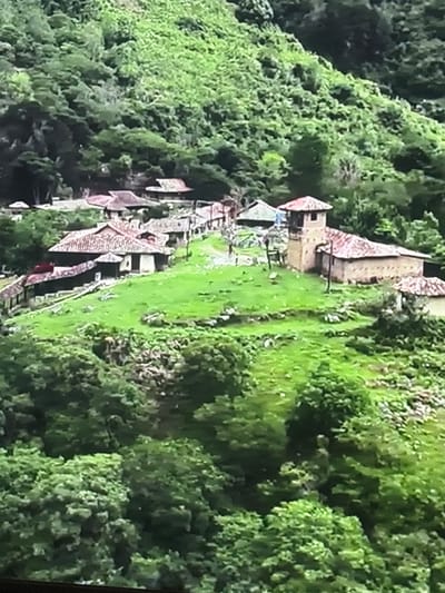 This is an aerial, slightly blurry, eye-level photograph of a rural village nestled into a lush, green hillside. The village consists of numerous buildings with red-tiled roofs, arranged in a cluster on a grassy slope. Some of the buildings are simple, single-story structures, while others are more complex, with courtyards and courtyards. One prominent building features a multi-story tower with a red-tiled roof, suggesting it might be a church or administrative building. The surrounding environment is densely forested with vibrant green trees covering the steep hillsides. The overall impression is one of a remote, traditional settlement integrated into a natural landscape. There are no people visible in the photograph. The image does not depict the city of Valladolid, Spain; rather, it appears to be a South American or European rural landscape.