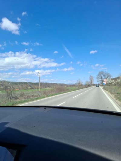 A roadside view from within a vehicle shows a gray asphalt road with white line markings extending into the distance. Signage is present. There is sparse vegetation on the right side of the road. Overhead utility lines are visible. The sky, predominantly blue, is populated with scattered white clouds. This image was taken in Komarevo, Bulgaria.