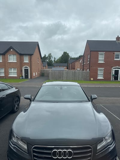The image depicts a residential street in Craigavon, United Kingdom, with two brick houses and their driveways visible. In the foreground, the hood and front grille of a dark grey Audi car are prominently displayed, taking up the lower half of the frame. To the left of the Audi, the rear section of a black car is visible, with its wheel and part of the bodywork showing. Both cars are parked on a paved surface. Behind the cars, a low wooden fence separates the properties from the houses. The houses are modern two-story dwellings constructed of red brick with dark tiled roofs and white-framed windows. The sky is overcast with grey clouds.