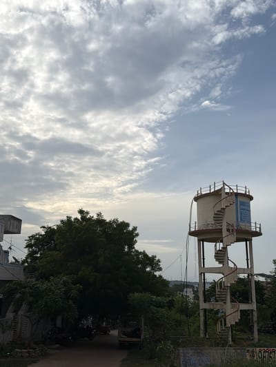 The image captures a view of a water tank with a spiral staircase on its exterior, located in the city of Chinna Tandrapadu, India. The water tank is a tall, cylindrical structure supported by pillars, painted in off-white with red accents around the top and bottom rims. A blue sign is affixed to the side of the tank. The spiral staircase, also in off-white with red railings, winds around the exterior of the tank.

In the foreground, a street or pathway is visible, lined with trees and some parked motorcycles. To the left, a building with a dark-colored facade and what appears to be a satellite dish is partially visible. The area is lush with vegetation, with large, dense trees dominating the mid-ground. In the background, more buildings and trees can be seen, suggesting a suburban or semi-urban environment. The sky is a prominent feature, filled with a mix of heavy, gray clouds and patches of brighter, lighter sky, indicating overcast or partly cloudy weather conditions. The overall scene depicts a typical day in a residential area of Chinna Tandrapadu, with the water tank serving as a significant landmark. There are no people or explicit actions depicted in the image.