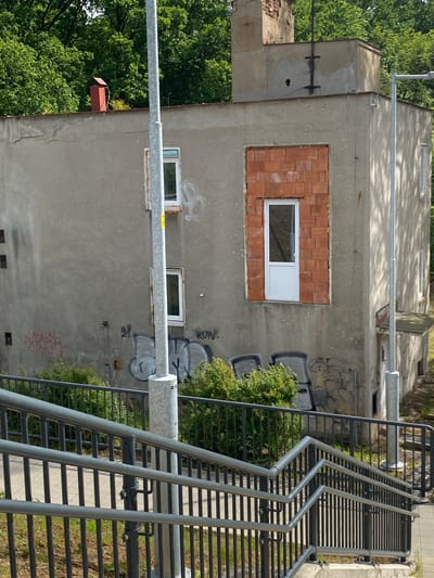 The image shows a three-story building in Lahošť, Czechia, with a grey concrete facade covered in graffiti. A section of the building has an unfinished brick opening with a white door. There are several small, dark square openings on the left side of the building, possibly former windows or ventilation shafts. A red chimney with a metal pipe extends from the roof. A metal staircase with handrails leads up to the building from the foreground. Trees and bushes surround the property. A light pole stands in front of the building, and another is visible on the right. The sky is bright and sunny.