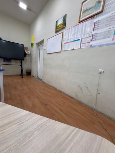 The image depicts an indoor scene, likely a classroom or office, with a light-colored wall and a wooden floor. On the wall, there are several items including a framed landscape painting, a smaller painting of a field, a whiteboard, and educational charts. A large television screen on a stand is visible in the left portion of the image, along with a blue and white table. Near the wall, there is a white electrical outlet with a cord plugged into it. The overall impression is of a functional space that may be used for educational purposes. There are no people present in the image. It is not possible to determine the specific location within Satovcha, Bulgaria from the visual information provided.