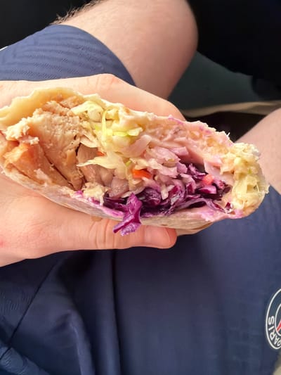 A person's hand holds a wrap filled with shredded chicken, coleslaw, and what appears to be red cabbage. The wrap is slightly open, revealing the colorful ingredients. The background is out of focus, but appears to be dark blue fabric, possibly clothing. No specific location in Hlybochytsia, Ukraine is identifiable.