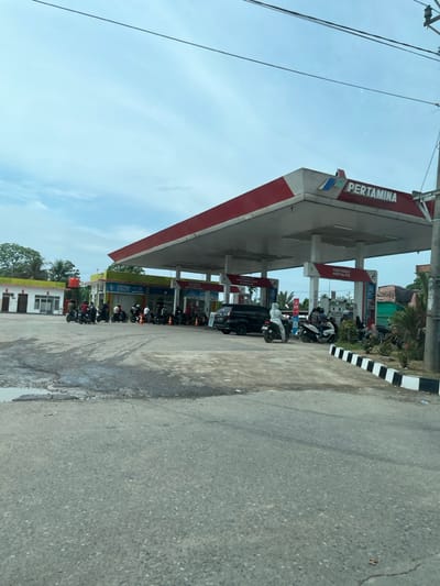 A Pertamina gas station stands under a cloudy sky. Motorcycles cluster near the pumps, some with drivers, others parked. The station's canopy has a red and white trim. Adjacent is a building with yellow accents. A black and white striped curb runs along the side of the road, which is cracked. Utility poles and wires are visible in the sky.
