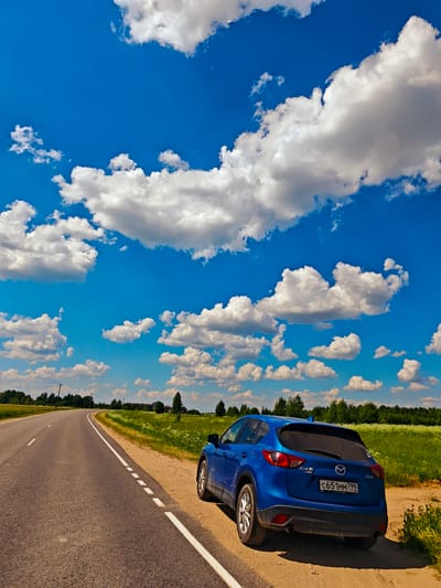 A blue Mazda CX-5 SUV is parked on the side of a rural asphalt road. The car is facing right, and its rear is visible. The road curves to the left and stretches into the distance. The environment is a bright, sunny day with a clear blue sky filled with fluffy white clouds. Fields of green grass and wildflowers line the road, with a dense line of trees in the background. Electrical poles and wires are also visible in the distance. The license plate on the car reads "C651MM 799 RUS". There are no people visible in the image. The scene appears to be a roadside in the countryside, possibly near the city of Kirovo, Russia.