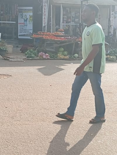 An adult male walks left-to-right across a paved street in Jebbu Miango, Nigeria. He wears a light green short-sleeved t-shirt with black side panels, blue jeans, and dark slide sandals, with an earphone cord visible on his left side. His elongated shadow extends diagonally forward on the light-colored asphalt. In the background, a bustling market scene unfolds along a storefront. A wooden stall displays numerous red tomatoes and possibly red onions. Below the stall, on the ground, are piles of green leafy vegetables, several large green watermelons, and smaller yellow and green produce. To the left, a "Breakforth Fashion House" sign is visible on a shop front, listing services such as "FASHION DESIGN," "TAILORING," and "READY TO WEAR CLOTHING." To the right, another storefront features a prominent vertical red sign reading "PHARMACY." The environment is brightly lit by direct sunlight, creating strong shadows.