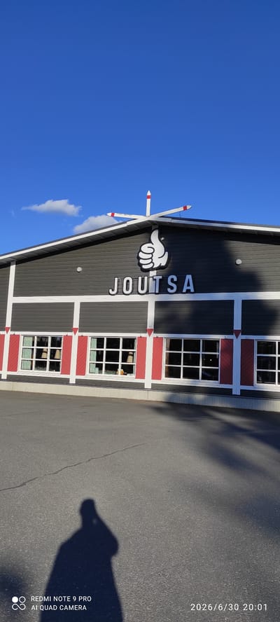 This is a rotated image of a building with the text "JOUTSA" displayed prominently on its facade. The building appears to be dark grey with white and red accents around windows and decorative elements. Above the "JOUTSA" text, there is a white cartoon-like hand pointing to the left. The building has multiple windows, each divided into a grid of smaller panes, and some red decorative panels below the windows. On the left side of the image, a bright blue sky with a few wispy clouds is visible. On the right side, a paved surface, likely a road or parking lot, is present with the shadow of a person or object casting a long, dark shape. The image also includes text overlay which reads "2026/6/30 20:01" and "REDMI NOTE 9 PRO AI QUAD CAMERA". The time stamp suggests the photo was taken in the evening. Given the text "JOUTSA" and the provided location context, it is highly probable that this building is located in Joutsa, Finland. The overall scene suggests an outdoor setting on a clear day, possibly late afternoon or early evening. There is no clear activity or event taking place, and no discernible emotions are conveyed.