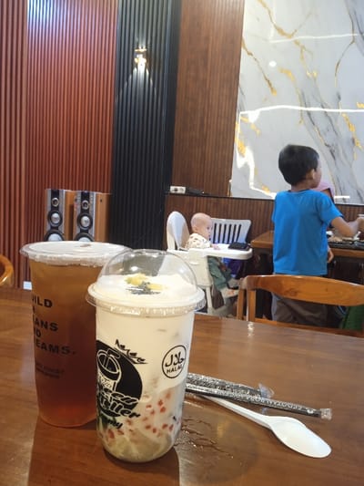 The scene depicts an indoor setting, presumably a cafe or restaurant, located in Pandean, Indonesia. The foreground features a polished wooden table surface where two beverages are placed. On the left, a tall transparent plastic cup contains a dark amber liquid, topped with a clear plastic lid. Visible text on this cup includes "BUILD YOUR DREAMS" and "TEA HOUSE". To its right, a shorter, wider transparent plastic cup holds a milky white liquid, incorporating red and green spherical components at the bottom. This cup is sealed with a domed clear plastic lid, adorned with whipped cream, dark seeds, and a slice of yellow fruit. A "HALAL" logo is distinctly printed on this cup. Next to this beverage are a black-wrapped straw, labeled "SEDOTAN BIODEGRADABLE", and a white plastic spoon. Minor condensation marks are visible on the table surface.
In the midground, a bald infant or toddler, clothed in light colors, is seated in a white high chair, facing left away from the viewer, with a white smartphone resting on the high chair's tray. To the right of the infant, the back of a young boy, wearing a blue t-shirt and grey shorts, is visible. He appears to be standing or sitting at a wooden table, looking towards the right side of the frame. A wooden chair back is partially visible in the lower right. The background wall is composed of several sections: the left segment features vertical reddish-brown wooden slats, a central part consists of dark vertical wooden slats with a small gold-colored wall sconce, and the right section displays a white marble pattern with prominent gold veining. A continuous lower panel of horizontal brown wooden slats runs across the base of these wall sections. Two silver-fronted audio speakers are positioned on a surface beneath the reddish-brown wall slats, to the left of the infant, and a power outlet is visible on the dark slat wall.