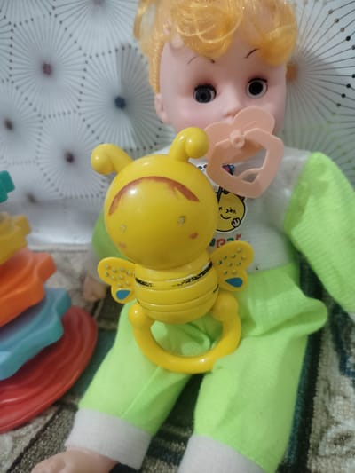 This is a close-up, eye-level, indoor photograph of a doll. The doll is a baby doll with blonde hair, blue eyes, and rosy cheeks. It is wearing a light-colored shirt with a cartoon bee graphic and a lime green outfit. The doll is holding a yellow bee-shaped rattle in its right hand, and a pink pacifier is in its mouth. A stack of colorful plastic stacking rings is visible to the left of the doll. The background consists of a patterned wallpaper with radial designs and a patterned rug underneath the doll. The image appears to be taken in a residential setting in Cholpon, Kyrgyzstan.