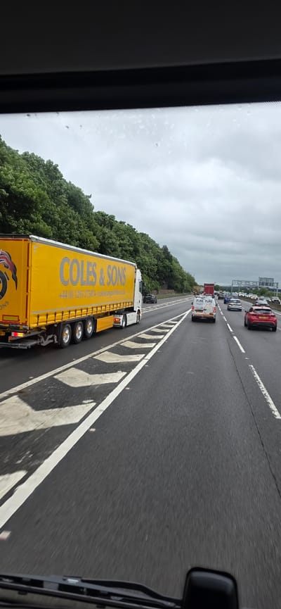 The image shows a view from inside a vehicle, looking out at a highway. A large yellow truck with "COLES & SONS" written on its side is prominently featured in the foreground, driving on the highway. Other vehicles, including cars and vans, are also visible on the road ahead. The highway is bordered by trees on the left and appears to be a multi-lane road. The sky is overcast. The scene is consistent with a view from a vehicle on a highway in the United Kingdom, possibly in or near Sheffield, given the general landscape and road markings.
