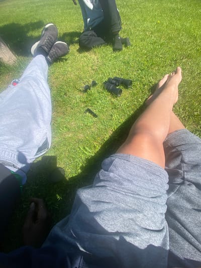 The image depicts a ground-level view of a sunny outdoor scene, presumably a park. Several individuals are present, with their legs and feet visible, suggesting they are sitting or lying down on the grass. One person is wearing dark sneakers and light grey sweatpants. Another individual's bare feet are visible, positioned near a pair of black binoculars. A third person, wearing dark pants and socks, is also visible. The environment is characterized by lush green grass, with some sparse yellow wildflowers. Shadows cast by an unseen object, possibly a tree or a structure, suggest the time of day is likely midday. There are no discernible landmarks indicating the specific location within Ajax, Canada.