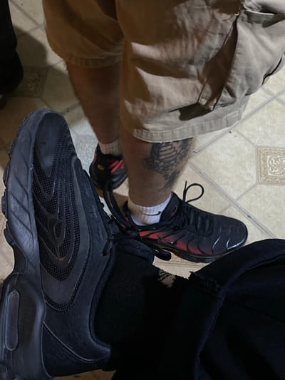 The image shows two pairs of legs and feet on a tiled floor. On the left, a person wears khaki shorts, white socks, and black sneakers with red accents. A tattoo is visible on the left leg. On the right, a person wears black pants, black socks, and black sneakers. The location, Monte Grande, Argentina, is not evident from the image.
