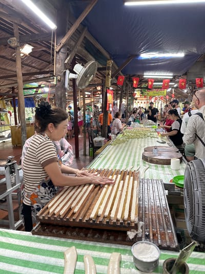 The image depicts an indoor scene in Tân Thạch, Vietnam, likely a market or workshop where coconut candy is being made. A woman in a striped shirt and floral apron is pressing a dough-like substance into a wooden mold with parallel grooves. The molds are arranged on a green and white striped surface where other candies are also present. Additional individuals, possibly customers or workers, can be seen further along the table. Vietnamese flags hang from the ceiling. Overhead are exposed wooden beams and fans. A security camera is also visible.
