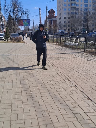 The scene depicts a young adult male walking on a grey paver sidewalk under a clear blue sky in Меткомелино, Russia. He is wearing a dark hooded puffer jacket, dark pants, and green sneakers, looking down at a smartphone held in his right hand; his left hand is in his pocket. A long shadow is cast to his left.
To the immediate right of the sidewalk, a black metal fence separates it from a road where several vehicles, including cars and vans, are parked or in motion. Patches of dirty snow are visible near the fence. Further right, a multi-story, light-colored residential building stands prominently, behind which a distinct red-brick building with a reddish-brown turreted roof and round windows is visible.
In the mid-ground to the left, several other pedestrians are walking on a similar sidewalk. A large billboard displaying an advertisement featuring a person in a green top holding a mobile phone is prominent. Bare trees and utility poles line the street. A tall, grey industrial smokestack is visible in the distant background on the left. Buildings of varying heights and styles are present on both sides of the street. The overall lighting suggests a bright, sunny day, likely in late winter or early spring given the bare trees and residual snow.
