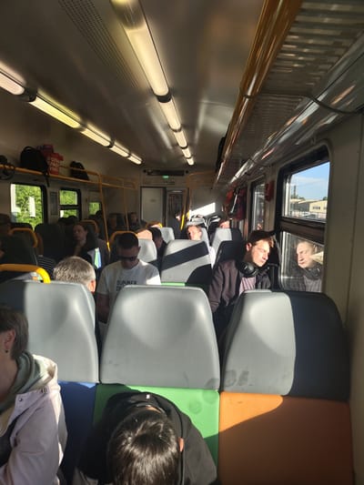 The image depicts the interior of a train carriage, viewed from the perspective of a seated passenger. Sunlight streams in through the windows, illuminating the aisle and casting long shadows. Several passengers are visible, some sleeping, others looking out the window or towards the front of the train. The seats are upholstered in grey with green and orange fabric accents on the lower sections. Luggage is stored on overhead racks. The train appears to be moving through a rural or suburban area, as indicated by the greenery visible through the windows. The number "06" is visible on a sign above a doorway. The scene is consistent with a commuter or regional train in Russia.